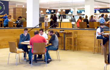 The inside of a common cafe area in the corporate offices where employees are gathered sitting at tables throughout to connect and enjoy lunch. 