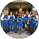 A diverse team of Bath and Body Works employees wearing their uniformed blue aprons holding a blue ribbon together in front of a new store location.